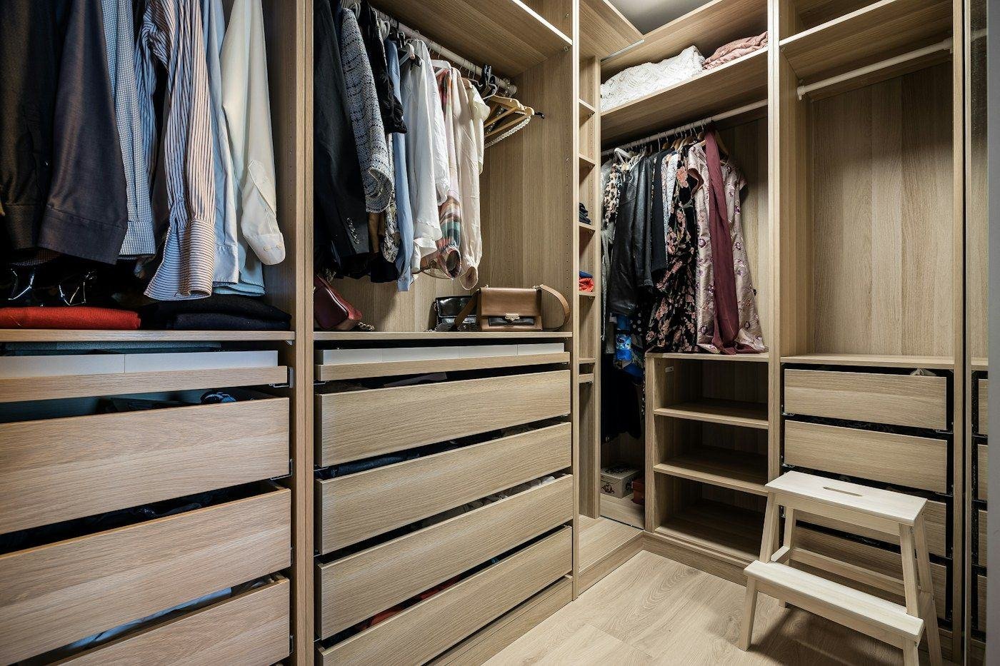 A fitted wardrobe with hanging clothes, wooden drawers, shelves and a small step stool.